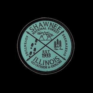 May include: A turquoise blue round patch with black stitching and text. The text reads "Shawnee National Forest" with a mountain range, pine trees, footprints, and arrows. The text "Est. 1933" and "Illinois Go Outside & Explore" are also included.