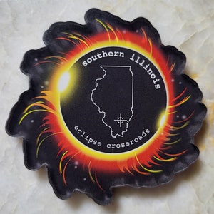 May include: A black and yellow sticker with a stylized image of the state of Illinois, surrounded by a ring of fire. The text "southern illinois eclipse crossroads" is written around the ring of fire.