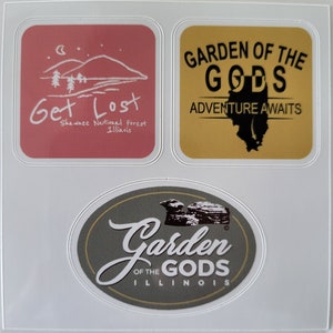 May include: Three stickers with a nature theme. The first sticker is pink with a white drawing of a mountain range and the text "Get Lost Shawnee National Forest Illinois". The second sticker is yellow with the text "GARDEN OF THE GODS ADVENTURE AWAITS" and a black silhouette of the state of Illinois. The third sticker is oval shaped with a gray background and the text "Garden of the Gods Illinois" in white with a brown silhouette of a rock formation.