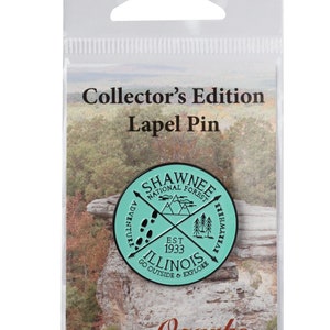 May include: A teal blue enamel pin with a black outline featuring the text "Shawnee National Forest" and "Illinois" with a compass design and the text "Go Outside & Explore" and "Est. 1933".