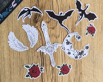 May include: A set of black and white stickers featuring dragons, a sword, flowers, and a crescent moon. The stickers are on a wooden surface.