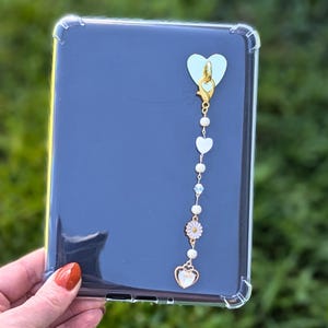 May include: A clear e-reader case with a decorative charm. The charm includes a light green heart-shaped adhesive, a gold-coloured clasp, white heart-shaped beads, and a daisy charm. The e-reader is dark blue.