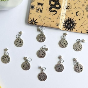 Könnte beinhalten: Ein Set aus zehn silbernen Charms mit verschiedenen Sternzeichen-Konstellationen. Jeder Charm ist ein Kreis mit schwarzem Umriss und einem Sternmuster. Die Charms sind an silbernen Ketten mit einem Stern am Ende befestigt.