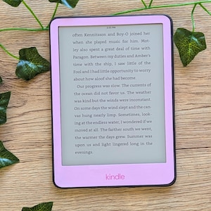 Peut inclure: Une liseuse Kindle rose avec un écran blanc affichant du texte d'un livre. Le texte dit : "souvent Kennitsson et Boy-O la rejoignaient lorsqu'elle jouait de la musique pour lui. Motley passait également beaucoup de temps avec Paragon. Entre mes fonctions et le temps d'Amber sur le navire, j'ai peu vu le Fou et j'ai eu peu d'occasions de me soucier de son détachement. Notre progression était lente. Les courants de l'océan ne nous étaient pas favorables. Le temps était clément mais les vents étaient inconstants. Certains jours, le vent dormait et la toile pendait presque mollement. Parfois, en regardant l'eau infinie, je me demandais si nous nous déplacions. Plus nous allions vers le sud, plus les journées étaient chaudes. L'été était sur nous et la lumière persistait longtemps dans les soirées."