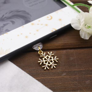 May include: A gold-colored snowflake charm with clear crystal accents hangs from a phone. The charm is attached to the phone with a clear, plastic piece. The phone is black and has a white and gold patterned case. The background is a dark wood surface.