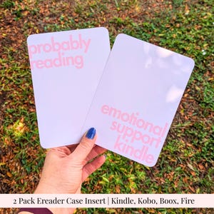 May include: A two-pack of rectangular ereader case inserts with rounded corners. One white insert features the pink text "probably reading" and the other white insert features the pink text "emotional support kindle" in a bold sans-serif font.