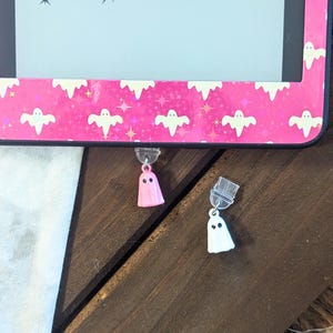 Puede incluir: Dos colgantes con forma de fantasma, uno rosa y otro blanco, cuelgan de unos enganches transparentes. Los colgantes tienen ojos de puntos negros. El fondo incluye un marco rosa con patrones de fantasmas y estrellas blancas, lo que sugiere un tema de Halloween.