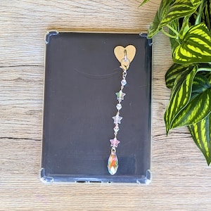 May include: A black tablet in a clear case with a gold heart charm attached. A beaded chain of iridescent stars and a teardrop-shaped crystal hangs from the heart. The background is a light wood surface with a green plant.