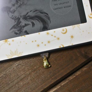 May include: A gold-colored owl charm attached to a clear plastic clip, hanging from the edge of a white e-reader case. The case has a celestial design with gold suns, stars, and constellations. The e-reader displays text that reads "100 Greatest Fantasy Books."