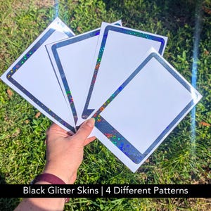 May include: Four white rectangular stickers with black glitter borders and holographic accents are held against a backdrop of green grass. The stickers have a rectangular shape with rounded corners. The text "Black Glitter Skins | 4 Different Patterns" is at the bottom.