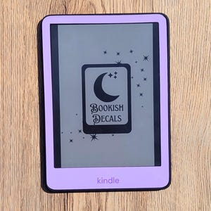 May include: A lavender Kindle e-reader with a black screen displaying a decal. The decal features a crescent moon, stars, and the text "Bookish Decals." The word "kindle" is printed at the bottom of the device.