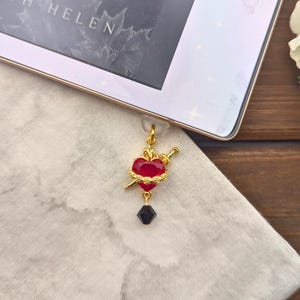 May include: A gold charm with a red heart pierced by a sword and a black crystal hanging from it.