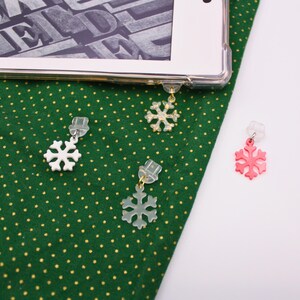 May include: Close-up of snowflake-shaped earrings in white, gold, and pink. The earrings are attached to clear plastic clips. The background is a green fabric with gold polka dots and a white surface.