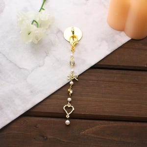 May include: Gold-colored charm with a lobster clasp, adorned with a series of decorative elements. These include a rhinestone-studded heart, a bow, and a dangling heart-shaped pendant, all connected by small pearls. The charm is designed for use as a keychain or accessory.