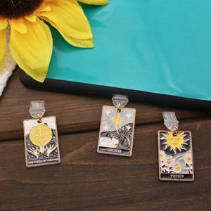 Puede incluir: Tres clips de marcador de oro con fondo negro y detalles blancos y amarillos. Los clips de marcador presentan imágenes de cartas de tarot: La Rueda de la Fortuna, La Estrella y El Sol.