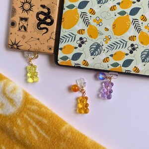 May include: Two yellow gummy bear charms and one purple gummy bear charm hanging from gold chains. The charms are attached to clear plastic clips. The clips are attached to two different book covers. One book cover has a lemon and bee pattern. The other book cover has a gold and brown pattern with a snake and an hourglass.