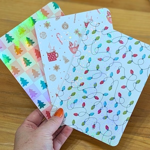 Può includere: Tre biglietti decorativi a tema natalizio con accenti olografici. Un biglietto presenta alberi di Natale, un altro ha tazze, bastoncini di zucchero e fiocchi di neve, e il terzo mostra luci di stringa. I biglietti hanno angoli arrotondati e una finitura lucida.