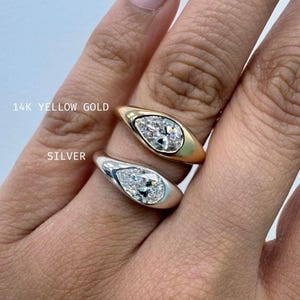 May include: Two rings featuring pear-shaped stones set in smooth metal bands. The top ring is labeled 14K YELLOW GOLD and the bottom ring is labeled SILVER. Both rings have a bezel setting that surrounds the faceted clear stone in a teardrop shape.