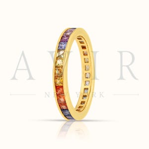 May include: A gold ring with a band of colorful gemstones. The gemstones are arranged in a rainbow pattern, with red, orange, yellow, green, blue, and purple hues. The ring is set against a white background.