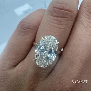 May include: A ring featuring a large oval-cut clear gemstone set in a gold-toned metal band with four prongs. The stone is faceted and reflects light. The text 6 CARAT is printed in the bottom right corner of the image.