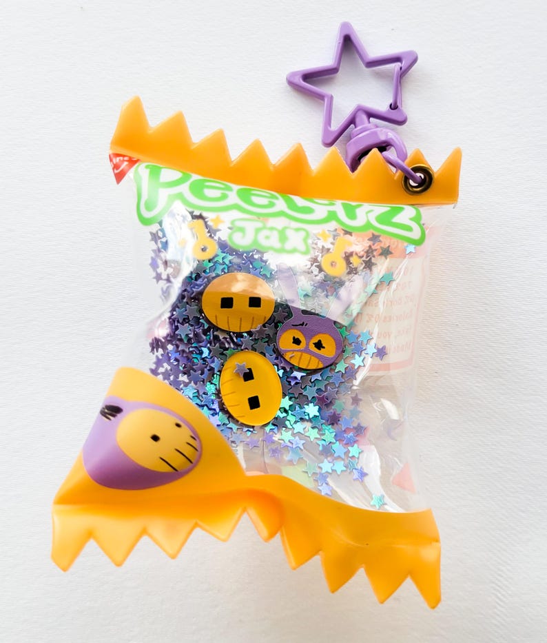 Może przedstawiać: Przezroczysta plastikowa torba z napisem "Peeps Jax" zawiera fioletowe konfetti w kształcie gwiazd i trzy ozdobne guziki. Torba ma pomarańczową, ząbkowaną g&oacute;rę i fioletowy brelok w kształcie gwiazdy.