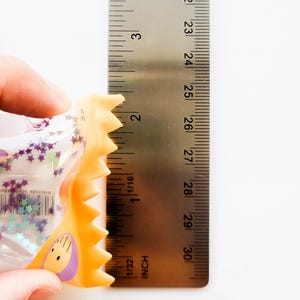 Może przedstawiać: Przezroczysta plastikowa zabawka z fioletowymi gwiazdkami i ż&oacute;łto-pomarańczową obw&oacute;dką trzymana jest obok linijki. Zabawka ma około 5 cm długości. Linijka jest oznaczona w calach.