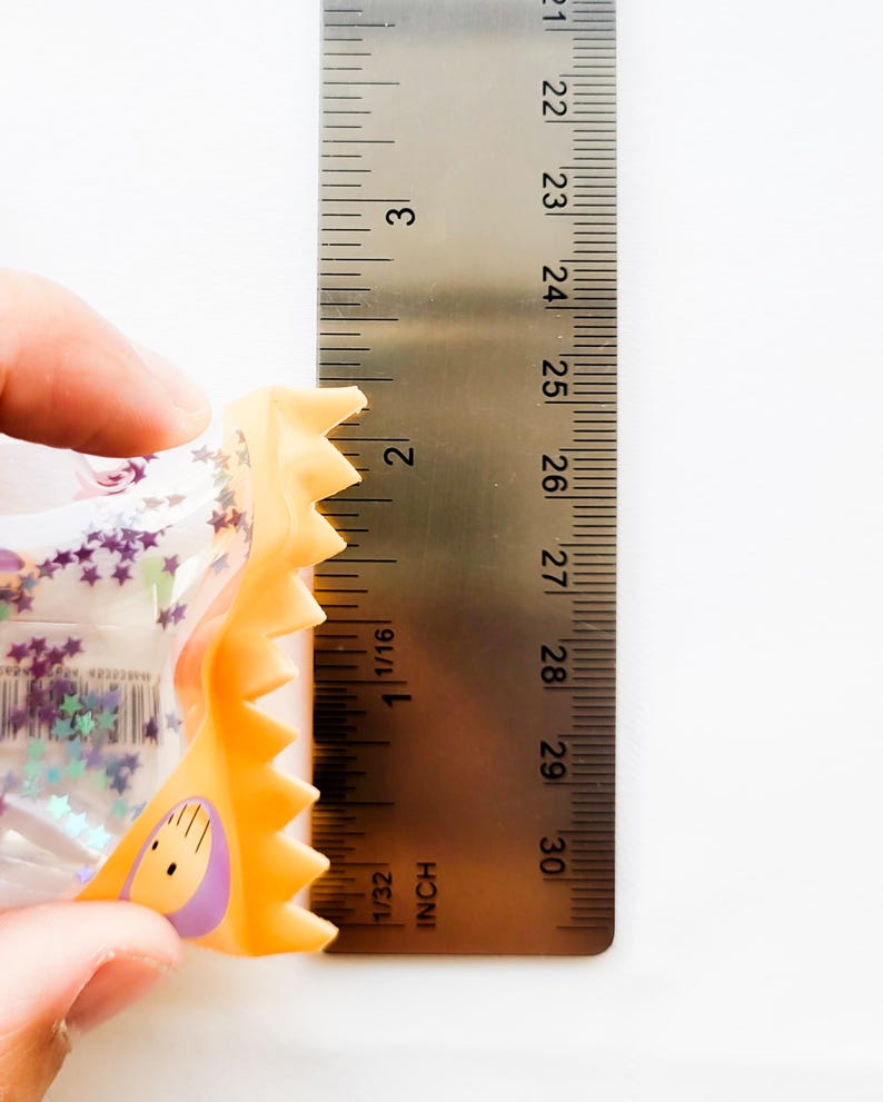 Może przedstawiać: Przezroczysta plastikowa zabawka z fioletowymi gwiazdkami i ż&oacute;łto-pomarańczową obw&oacute;dką trzymana jest obok linijki. Zabawka ma około 5 cm długości. Linijka jest oznaczona w calach.
