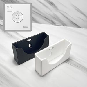 Könnte beinhalten: Zwei Wandhalterungen für eine Fernbedienung. Eine ist schwarz und die andere ist weiß. Beide haben ein abgerundetes Design, um die Fernbedienung zu halten.