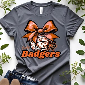 Può includere: Maglietta grigia con un design di pallavolo con un grande fiocco arancione e dettagli animalier. Il testo "Lady Badgers" è arancione, con un logo "KC". La maglietta è mostrata con jeans blu e scarpe color cuoio.