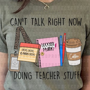 May include: An olive green t-shirt with the text "CAN'T TALK RIGHT NOW DOING TEACHER STUFF". The graphic features a lanyard, notepad, pens, and a coffee cup, all related to teaching.