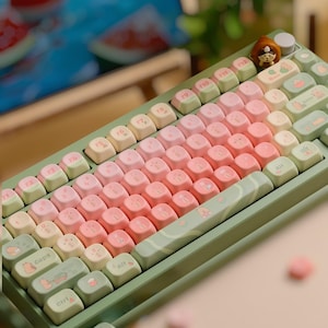 Puede incluir: Un teclado mecánico de colores pastel con un degradado de teclas rosas, melocotón y crema. El teclado tiene un marco verde y teclas decorativas con ilustraciones de dibujos animados. Una pequeña figura se encuentra en la esquina superior derecha.
