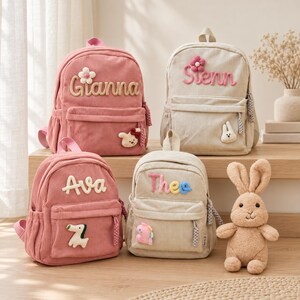 May include: Four corduroy backpacks in pink and beige, each featuring a personalized name in raised lettering, a small floral or animal-shaped patch, and a zippered front pocket. A plush tan rabbit toy sits beside the backpacks.