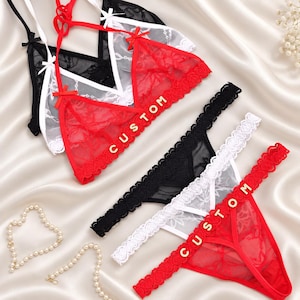 Pode incluir: Um conjunto de três bralettes e tangas de renda em preto, branco e vermelho. Cada peça apresenta renda floral transparente, laços delicados e letras douradas que formam a palavra CUSTOM nos itens vermelhos. O conjunto de lingerie é exibido.