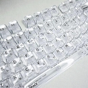 May include: Clear keyboard cover with black lettering. The cover is transparent, allowing the keyboard keys to be visible. The keys have English and Korean characters. The space bar has the text "Switch to hand tool (hold) SPACE".