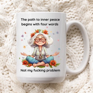 May include: White ceramic mug with a cartoon illustration of a woman meditating with flowers in her hair. The text reads "The path to inner peace begins with four words: Not my fucking problem."