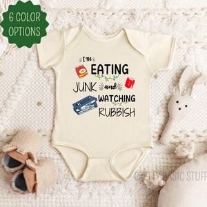 May include: A cream-colored baby onesie with the text "I'm eating junk and watching rubbish" in black. The onesie features a red bag of chips, a red can of soda, and a VHS tape.