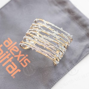 Puede incluir: Brazalete de puño dorado con un diseño en capas. El brazalete presenta múltiples barras horizontales con una apariencia texturizada en forma de rama, adornadas con cristales brillantes. El brazalete descansa sobre una bolsa de tela gris con la marca "alexis bittar" en naranja.