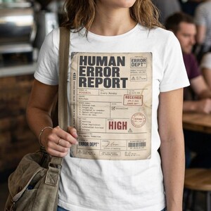 Peut inclure: Un t-shirt blanc à manches courtes avec une grande impression rectangulaire ressemblant à un document vintage intitulé Human Error Report. Le motif comprend du texte noir, un tampon rouge indiquant High, un tampon Received et des taches de café.