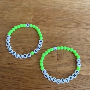 Puede incluir: Dos pulseras de cuentas verde neón. Cada pulsera presenta cuentas de letras blancas que deletrean "LANDOSTAND" y números. Una pulsera tiene "2025" y la otra "2026". Las pulseras están sobre una superficie de madera.