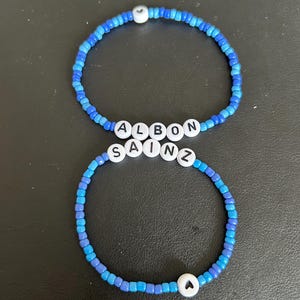 Könnte beinhalten: Zwei blaue und weiße Perlenarmbänder mit den Namen "Albon" und "Sainz" darauf. Jedes Armband hat einen kleinen weißen Herzanhänger.