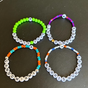 Könnte beinhalten: Vier Perlenarmbänder mit weißen Perlen, die "LANDO NORRIS 2025", "LANDO MIAMI", "NORRIS SINGAPORE" und "NORRIS HUNGARY" buchstabieren.