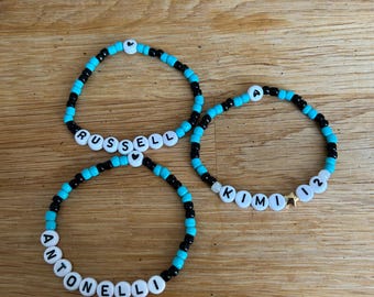Puede incluir: Tres pulseras circulares de cuentas con colores negro y turquesa alternados. Una pulsera muestra el nombre RUSSELL, otra el nombre ANTONELLO y la tercera presenta el nombre KIMI seguido del número 02 y una estrella dorada.