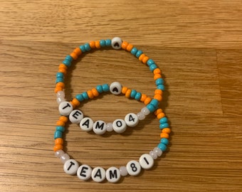 May include: Two beaded bracelets with the words "TEAM 04" and "TEAM 81" spelled out in white letter beads. The bracelets feature orange, blue, and white beads. The bracelets are on a wooden surface.