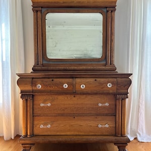 May include: A vintage wooden dresser with a mirror on top. The dresser has four drawers with crystal knobs and two long, silver handles. The dresser is a rich brown color and has a carved design on the sides. The dresser is sitting on a wooden floor.