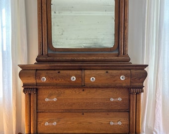 May include: A vintage wooden dresser with a mirror on top. The dresser has four drawers with crystal knobs and two long, silver handles. The dresser is a rich brown color and has a carved design on the sides. The dresser is sitting on a wooden floor.
