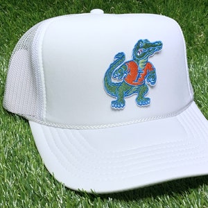 May include: White trucker hat with a green alligator wearing an orange shirt and a blue letter F on the front. The hat has a white mesh back.