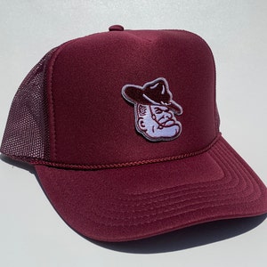 May include: Maroon trucker hat with a mesh back and a curved brim. The hat features a patch of a cartoon cowboy with a hat. The hat has a rope detail along the brim.