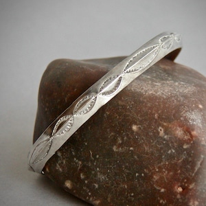 May include: A silver-toned metal bangle bracelet featuring a repeating pattern of elongated, pointed oval shapes with fine, etched hatch marks along the edges, creating a textured, geometric design across the surface of the band.