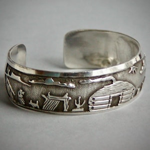 May include: A silver cuff bracelet featuring a textured, oxidized background with a raised relief design of a log cabin, a small structure, a cactus, and a figure. The polished silver edges frame the detailed scene, creating a high-contrast look.