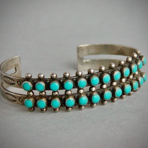 May include: A silver cuff bracelet featuring two parallel bands adorned with rows of small, round, bright turquoise stones. The metal surface displays decorative stamped leaf-like patterns near the open ends and small silver beads between stones.